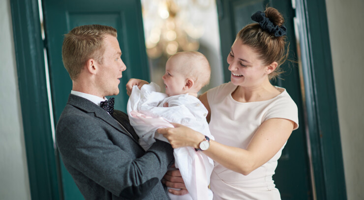 Far og baby smiler glade til hinanden, mens mor giver barnet dåbskjolen på lige før barnedåb i kirken