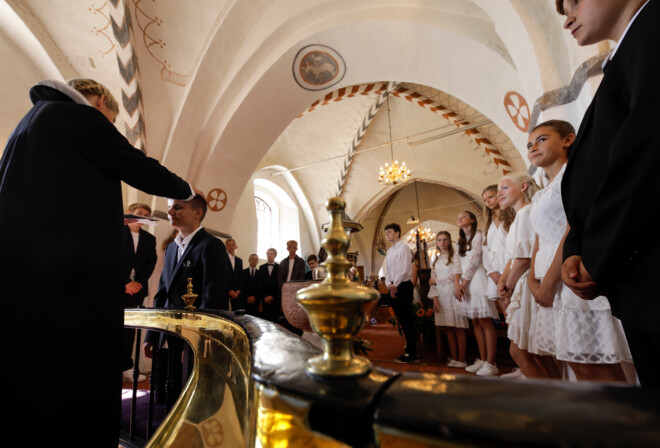 15 konfirmander er samlet foran alteret i en kirke, hvor præsten er i gang med at konfirmere en dreng ved at lægge hånden på hans hoved.